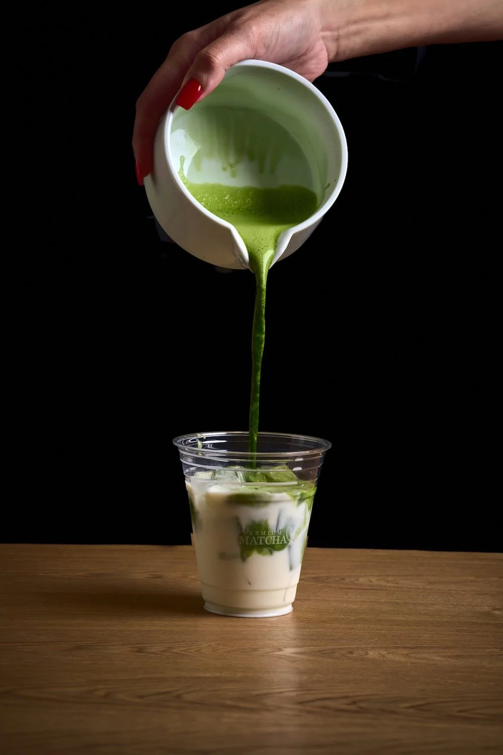 Matcha tea being poured from a white pitcher into a clear plastic cup with milk and matcha swirls inside, on a wooden surface against a black background at Matcha Cafe Maiko.
