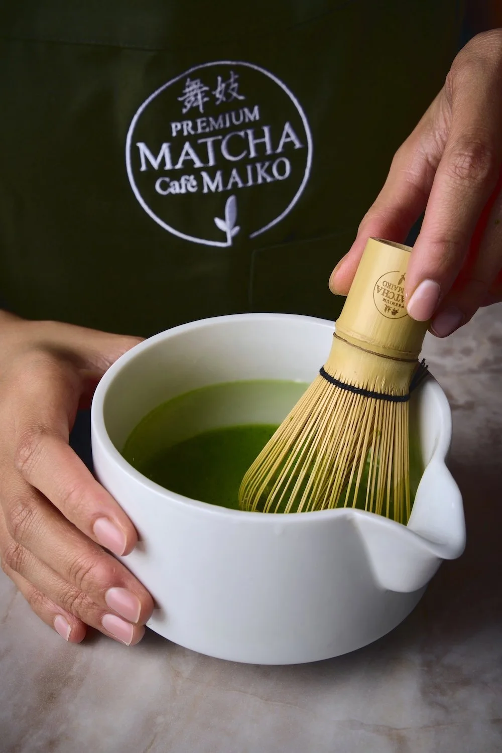 Person holds a cup of green matcha tea with a bamboo whisk inside, in front of a black shirt with white logo reading 'PREMIUM MATCHA Cafe MAIKO'.