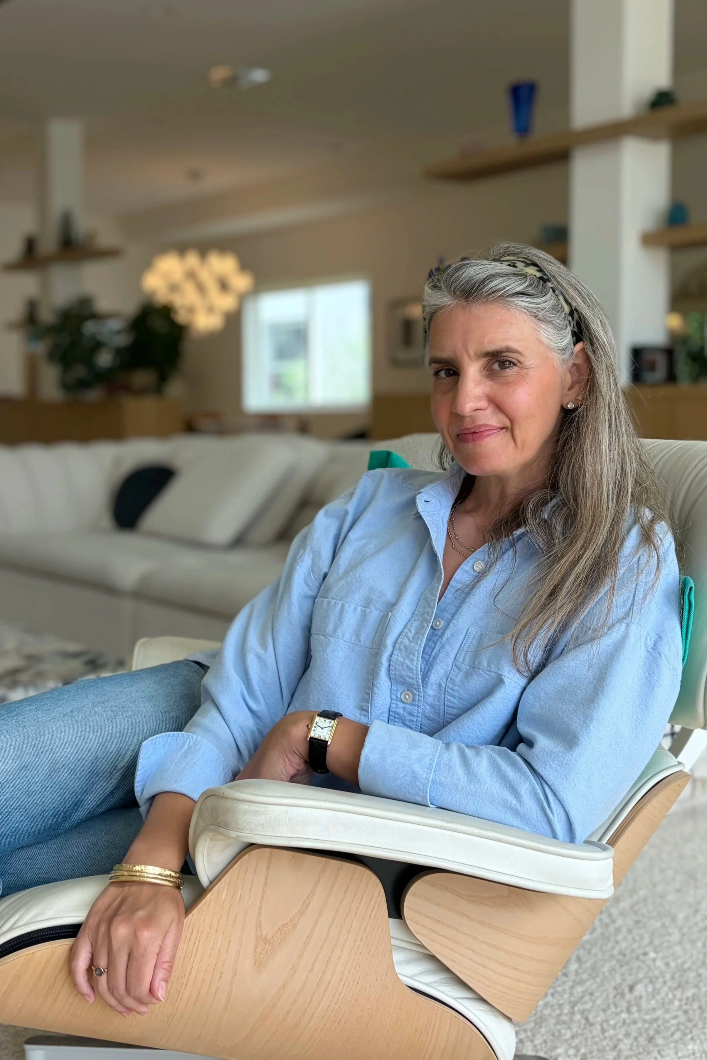 A woman with gray hair, wearing a headband, light blue shirt, and jewelry, sitting on a beige chair with a wooden tray attached, in a cozy room with shelves, plants, and blurred background.