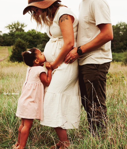 A woman with a hat, a man, and a young girl hold hands in a grassy field, with the woman showing a pregnant belly.