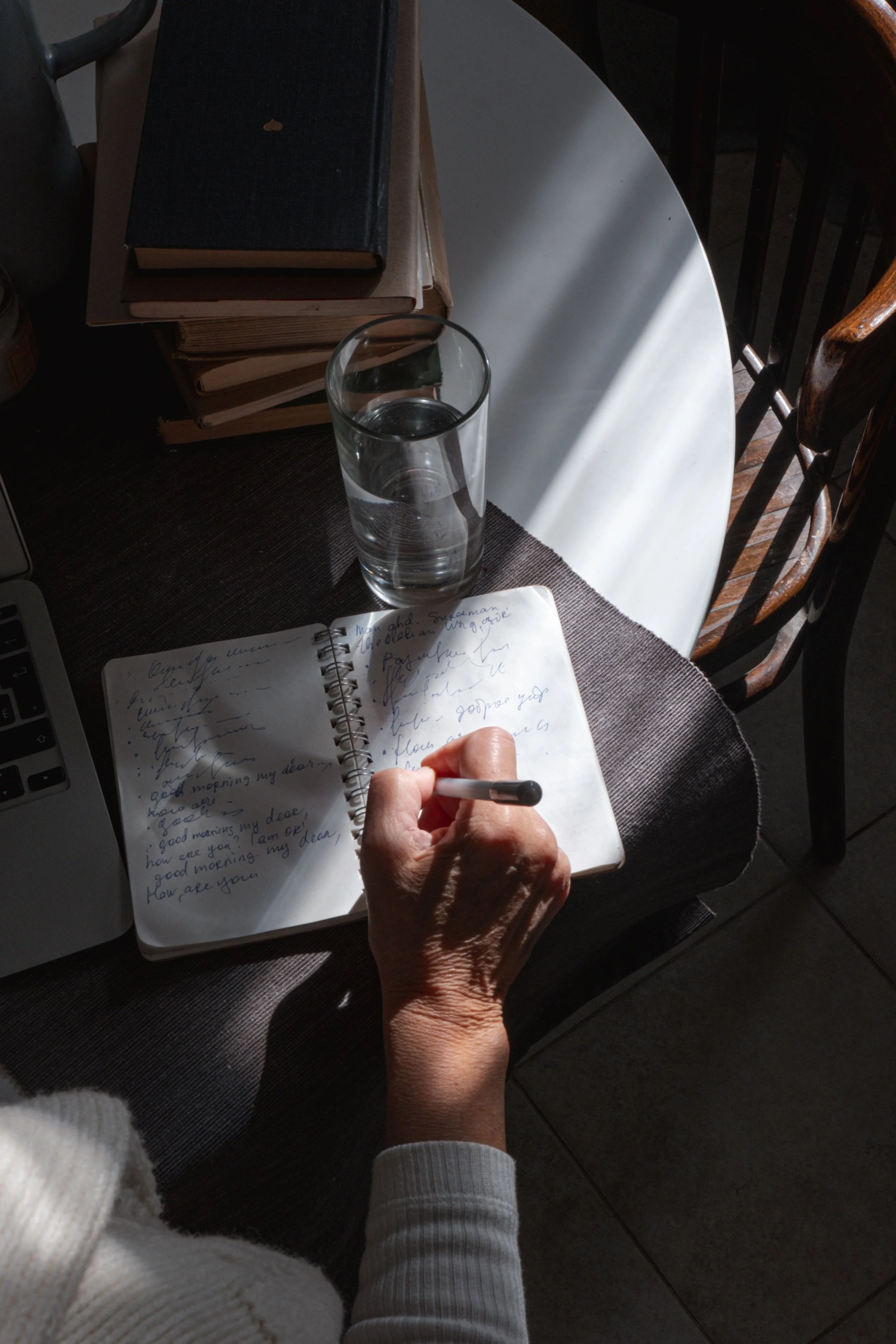 A person writing in a notebook on a round table with a glass of water, a stack of books, and a laptop nearby. The person is wearing a white sweater and has sunlit shadows on the table.