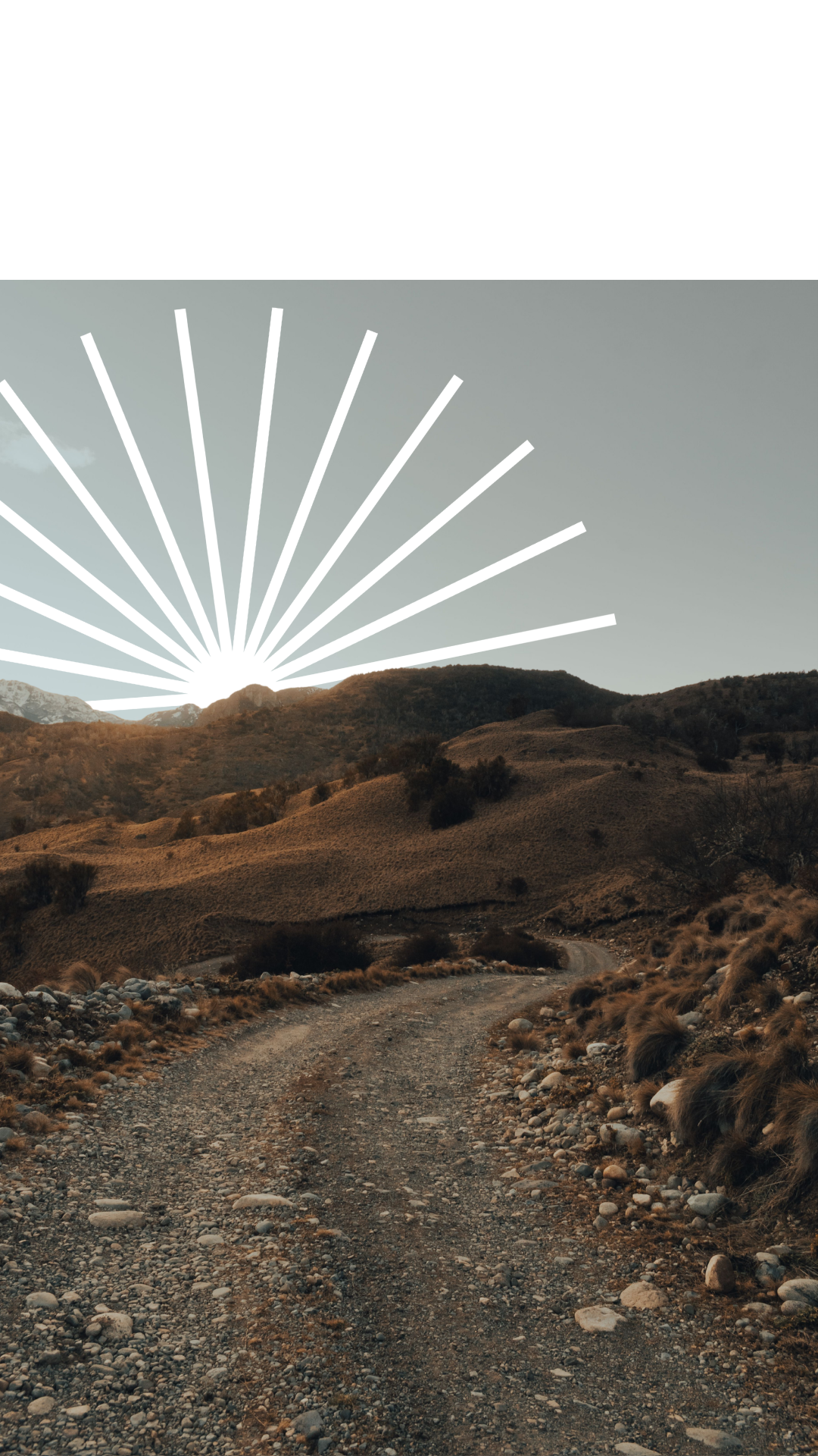 A rocky dirt road winding through hilly, brownish terrain with sparse trees and bushes, with the sun rising behind distant mountains and rays radiating upward.