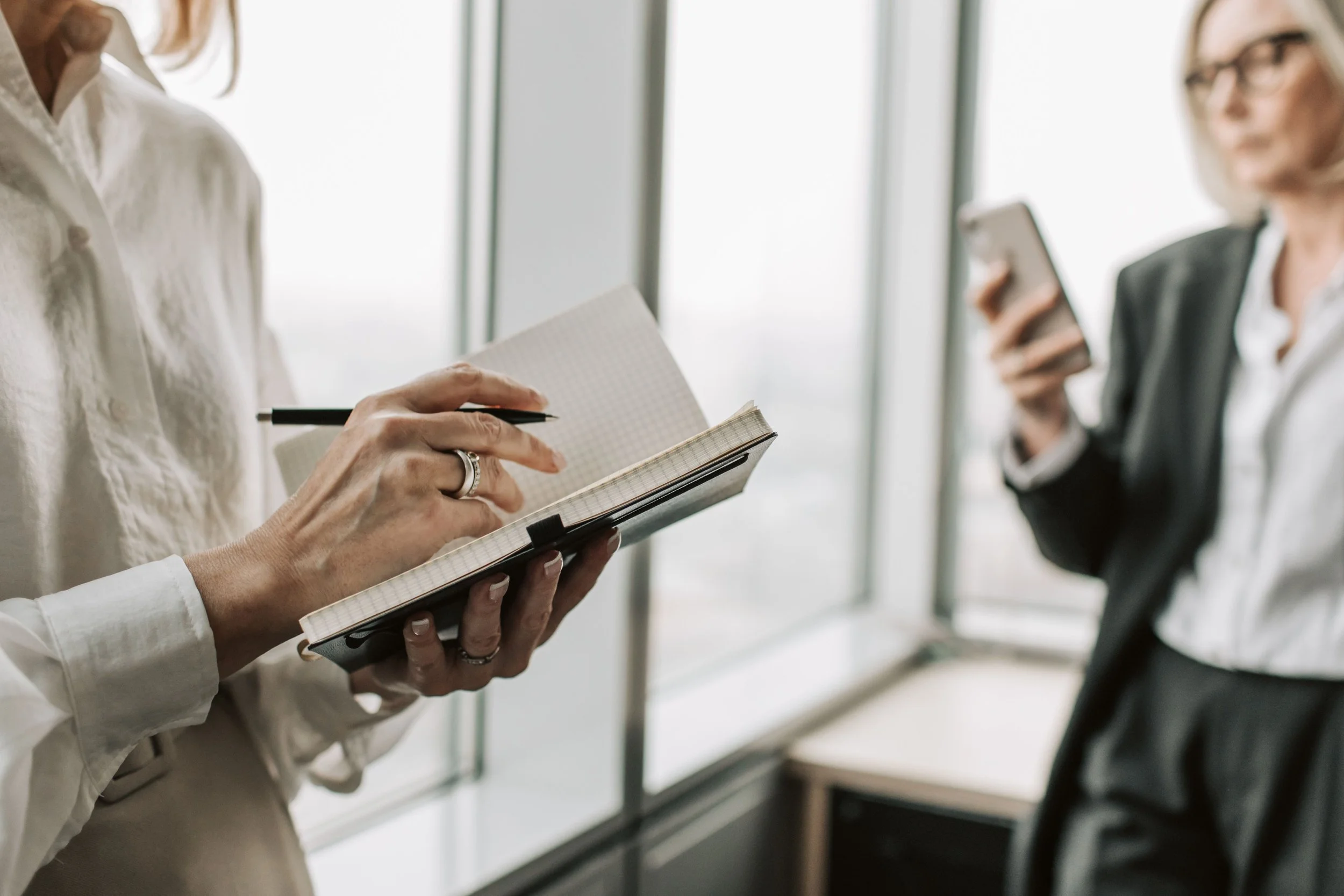 Two women engaged in a discussion, with one writing in a notebook and the other holding a smartphone, in a professional setting near large windows.