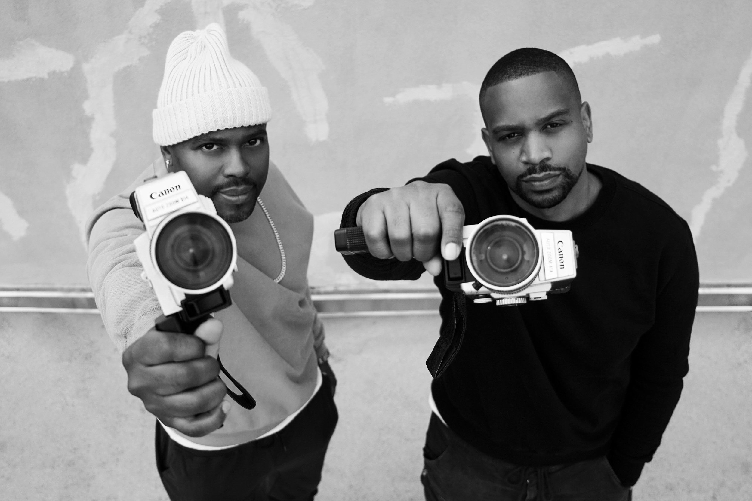 Two men standing close together, holding cameras pointed at the viewer, with serious expressions. One is wearing a white beanie and a sweatshirt, the other is in a dark shirt, against a textured wall background.