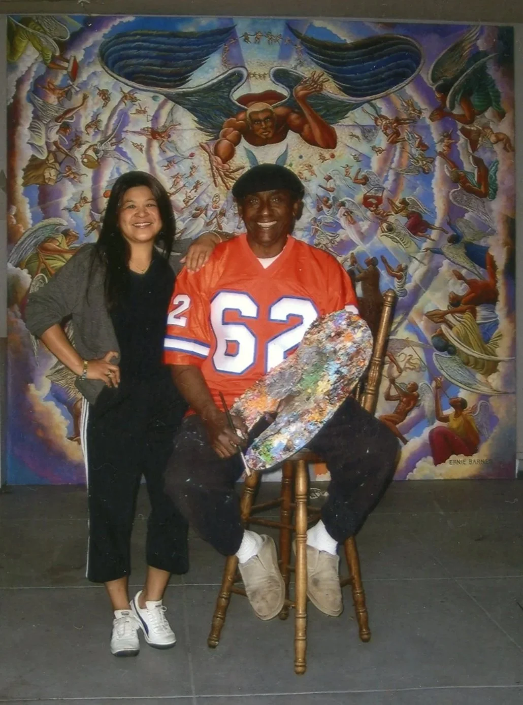 A woman and a man in a painted art studio with a large vibrant mural of angels and the sky in the background. The woman is standing to the left, smiling, with her arm resting on the man's shoulder. The man, sitting on a wooden chair and holding a painter's palette and brushes, is wearing an orange sports jersey with the number 62.