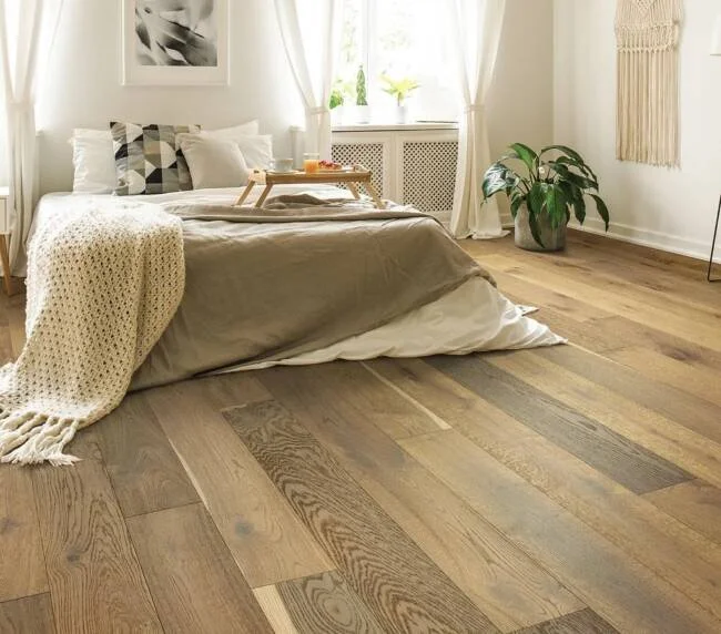A cozy bedroom with a low bed covered in beige bedding, a wooden tray with a drink on it, and a cream knitted blanket draped over the side. There is a large potted plant near the window, sheer white curtains, and wooden flooring.