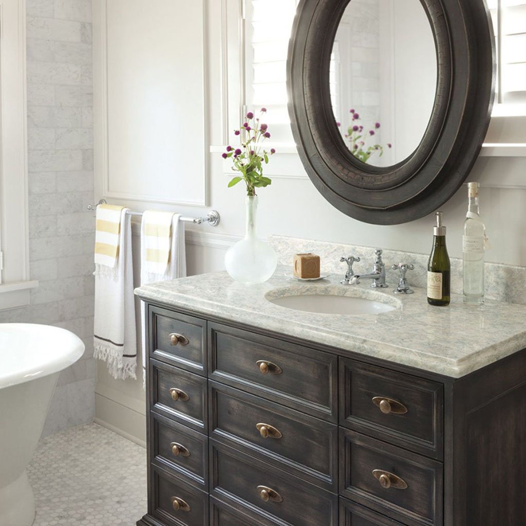 A bathroom featuring a dark wood vanity with a marble countertop, a round mirror, a vase with purple flowers, a soap, and bottles, and a window with white shutters.