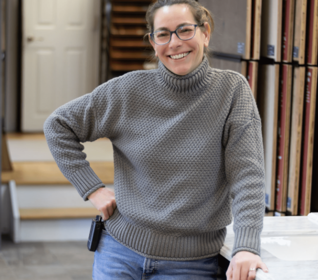A woman with glasses and a gray sweater smiling and leaning on a counter in a home or office setting.