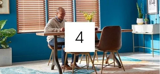An elderly man sitting at a dining table with three chairs in a brightly decorated room with blue walls, a window with blinds, and houseplants.