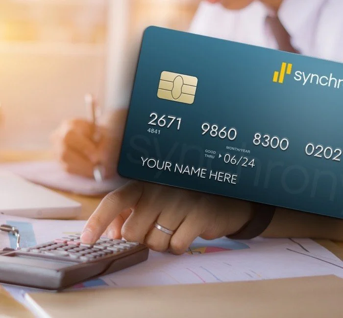 Close-up of a person holding a blue Synchron credit card with a chip, with a calculator and papers on a table.
