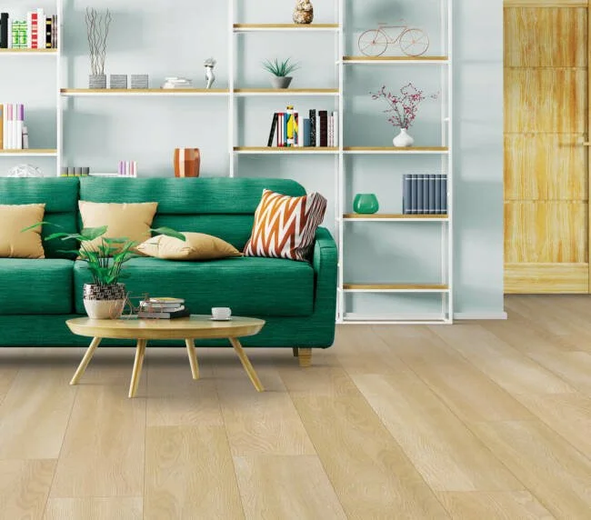 Living room with a green sofa, beige and patterned pillows, a light wood coffee table with a plant and magazines, white wall-mounted shelves with decorative items, and light wood flooring.
