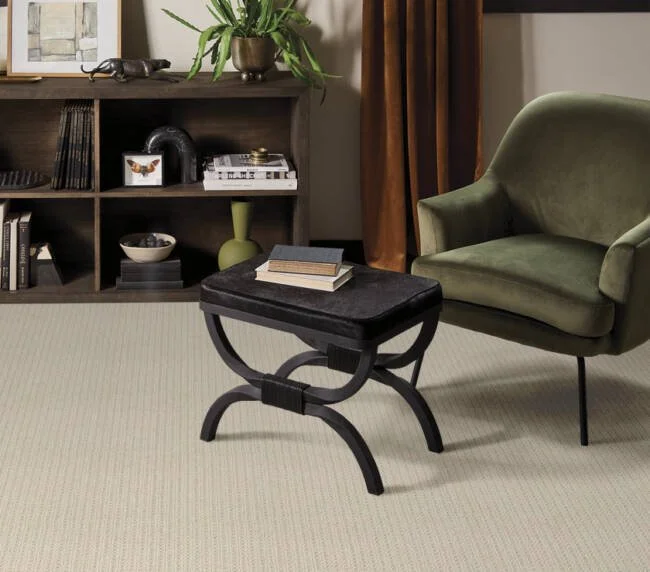 A side table with a black cushioned top holding some books, positioned next to a green upholstered armchair in a living room. There is a wooden shelf with decor items and books in the background, beige carpet on the floor, and a window with curtains.