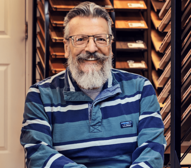 A smiling man with glasses and a beard, wearing a striped blue and gray sweater, sitting in front of shelves filled with mail or packages.