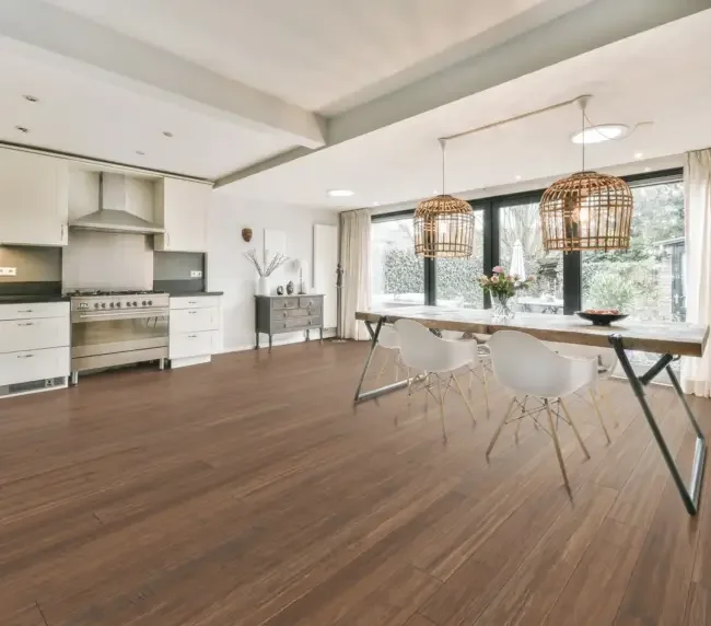 Empty dining area in a modern kitchen with large windows, a wooden table, white chairs, and woven pendant lights.