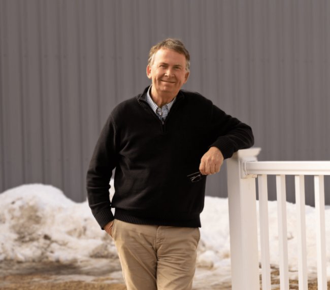 Smiling middle-aged man standing outdoors with snow, leaning on a white railing, wearing a black sweater and khaki pants.