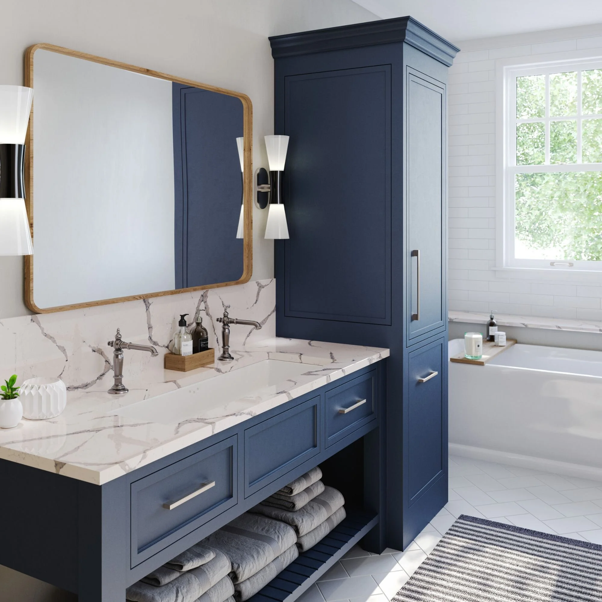 A bathroom with blue cabinetry and a white marble countertop with gray veining, two sinks, and a large mirror with a wooden frame. There are wall-mounted light fixtures with white shades, a window with white trim, and shelves underneath holding towels.