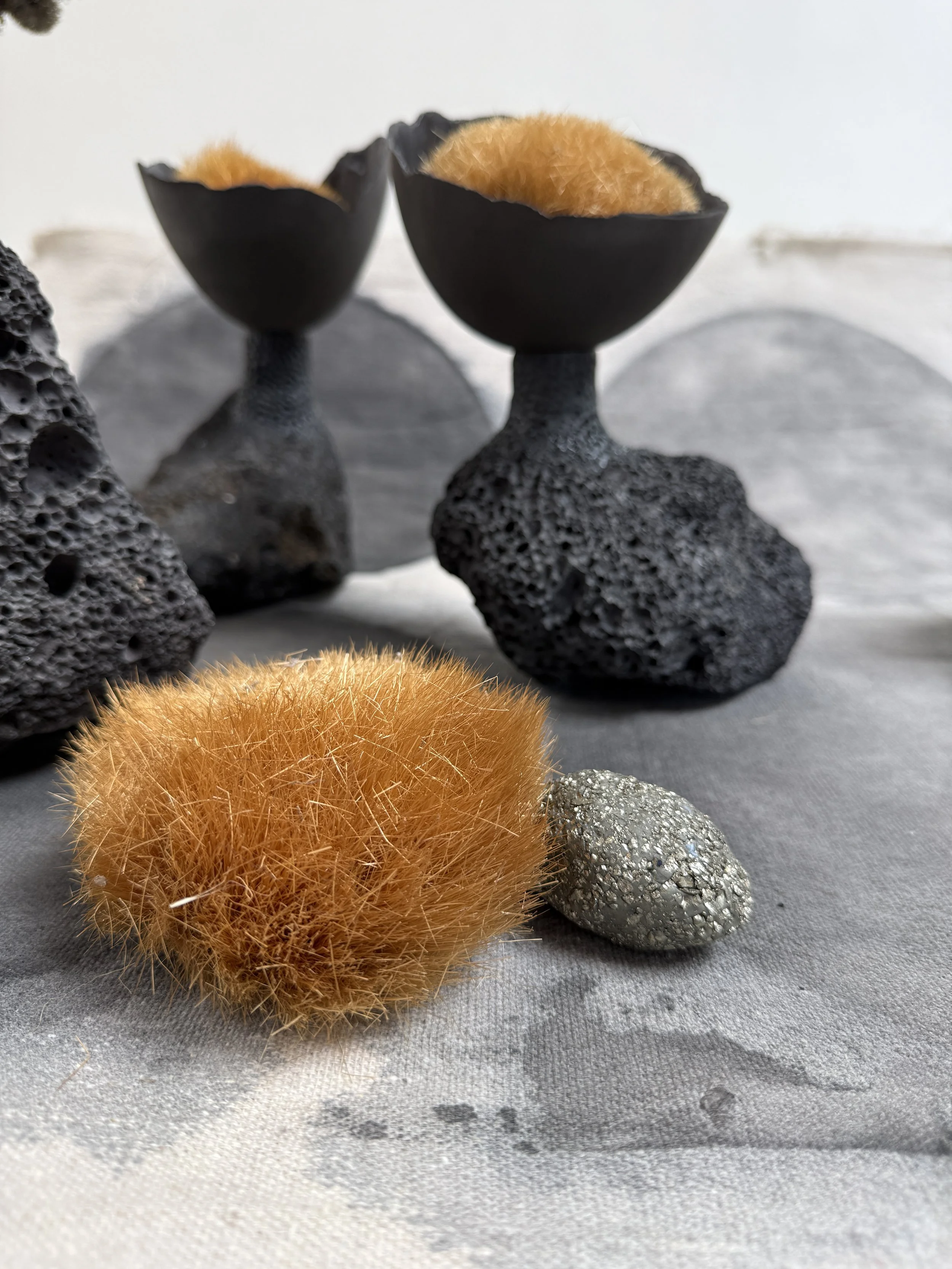Two black bowls with textured bases holding fluffy orange objects, surrounded by black volcanic rocks and shiny metallic rocks on a gray fabric surface.