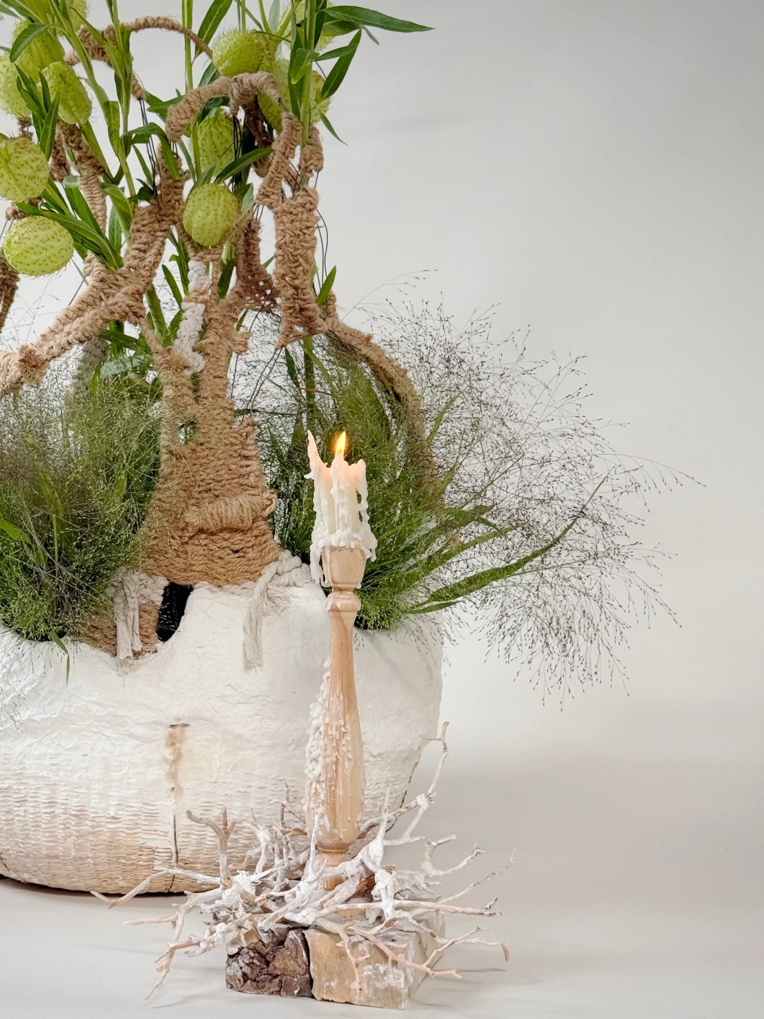 Woven Manzanita Candlestick by Kept LA styled in an interior  setting alongside fresh botanicals and the Weave and Plaster Basket,  showing lit taper candle with accumulated wax
