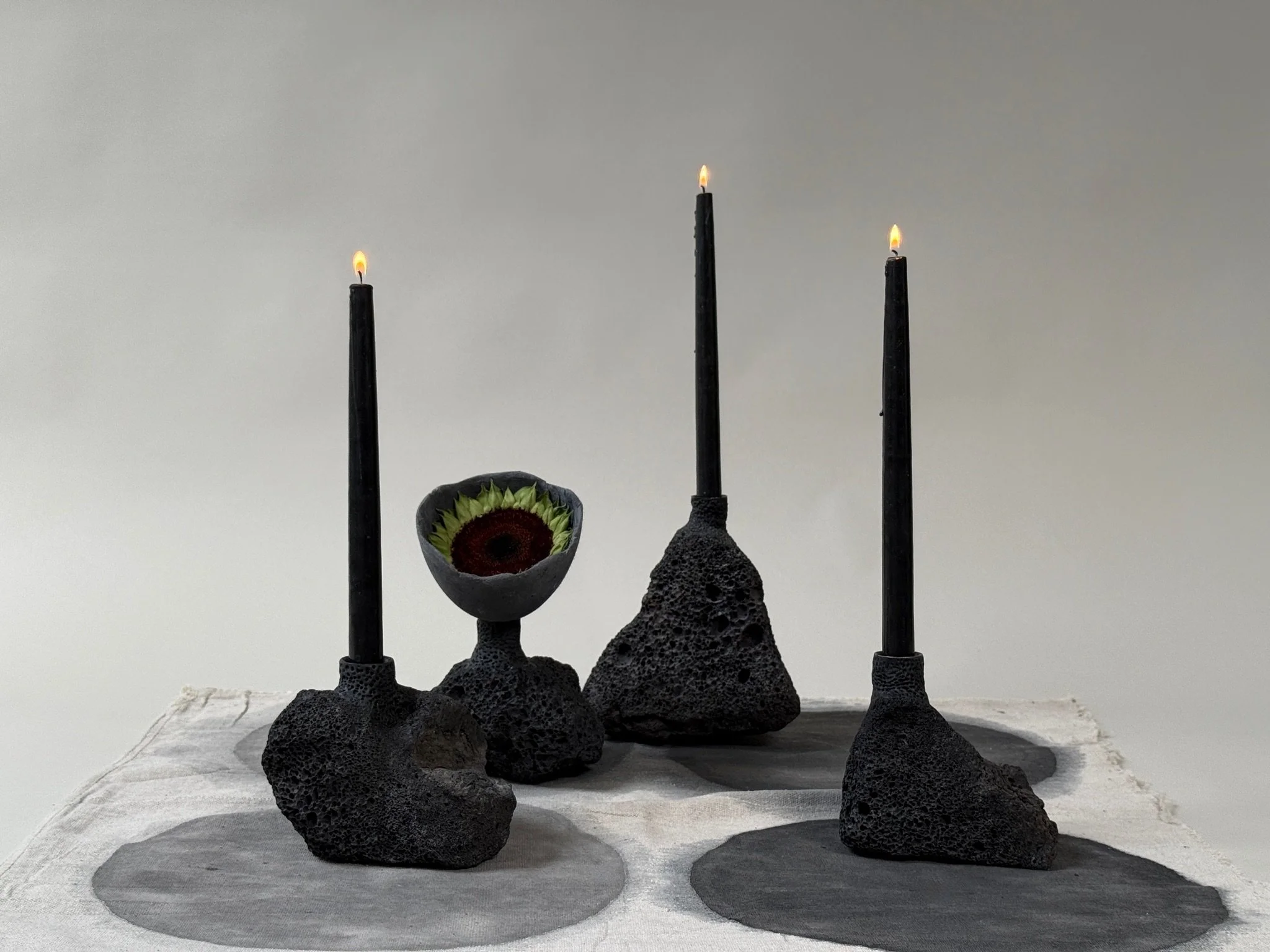 Volcanic Candlesticks by Kept LA with Little Cup inserts —  one holding a sunflower head, one with a lit black taper candle,  shown on hand-painted linen