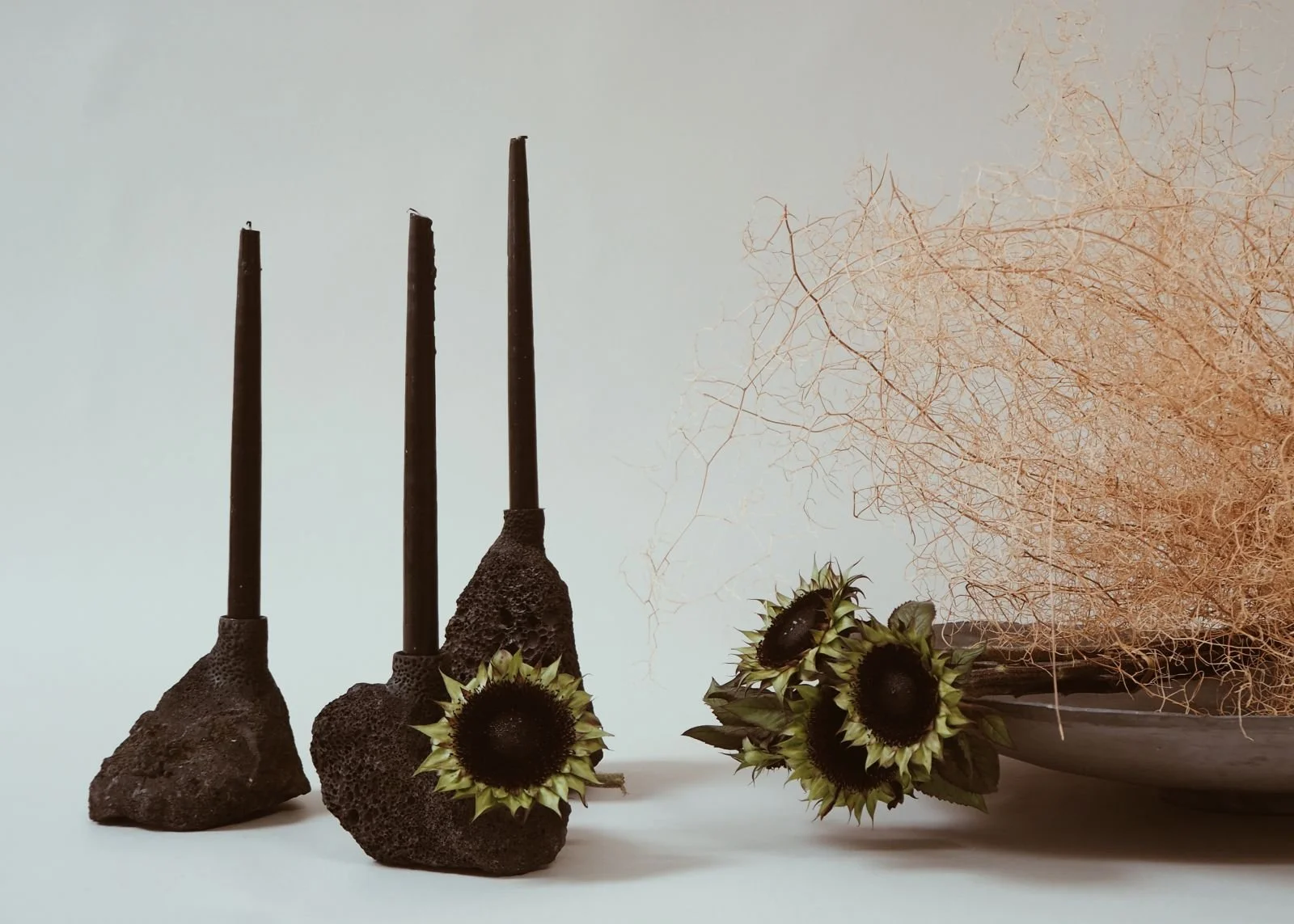 Decorative arrangement including three black candles in textured holders, dried branches, and sunflowers in a bowl.