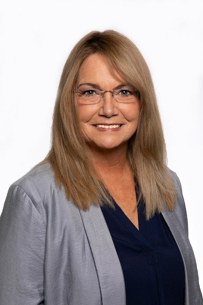 A middle-aged woman with blonde hair, wearing glasses, a dark blue blouse, and a light gray blazer, smiling against a white background.