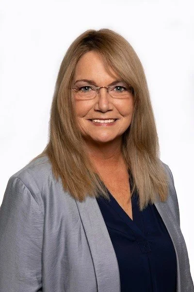 A middle-aged woman with blonde hair, glasses, and a friendly smile, wearing a navy blouse and a gray blazer, posing against a plain white background.