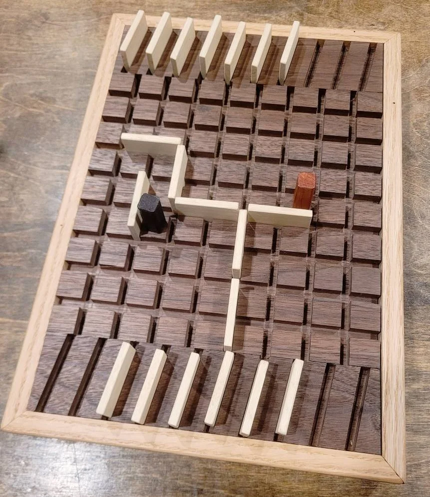 Quoridor.
Simple game of skill with 2 or 4 players. The goal is to reach the other side before your opponent by strategically placing walls and moving your piece. The board is made of walnut and the frame is red oak. I added a slide out drawer where 