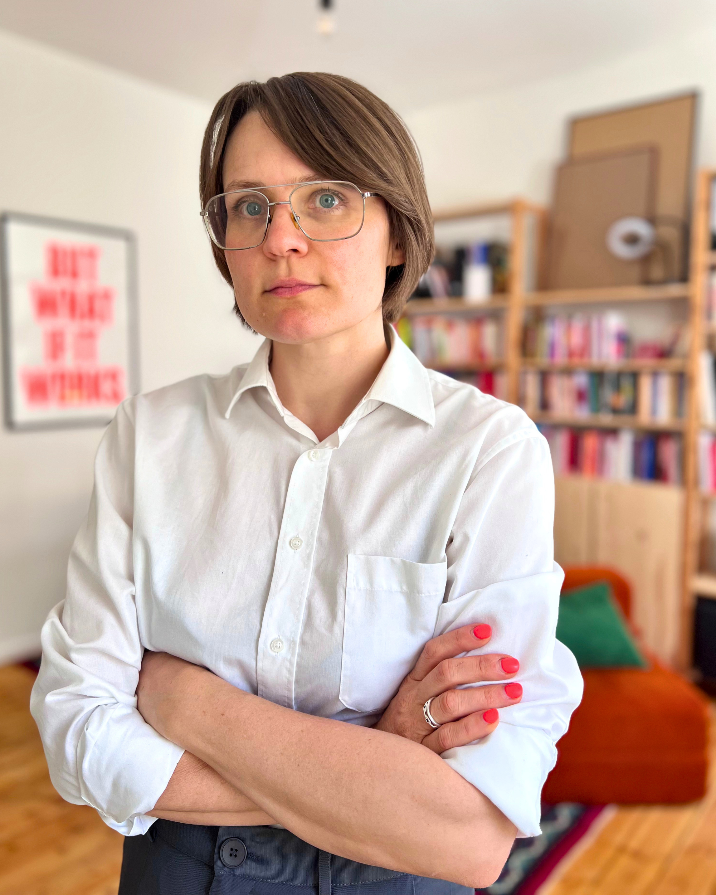 Eine Frau mit kurzen braunen Haaren, Brille mit dünnem Rahmen, weißem Hemd und dunklen Hosen steht mit verschränkten Armen in einem Raum mit einem Bücherregal im Hintergrund.