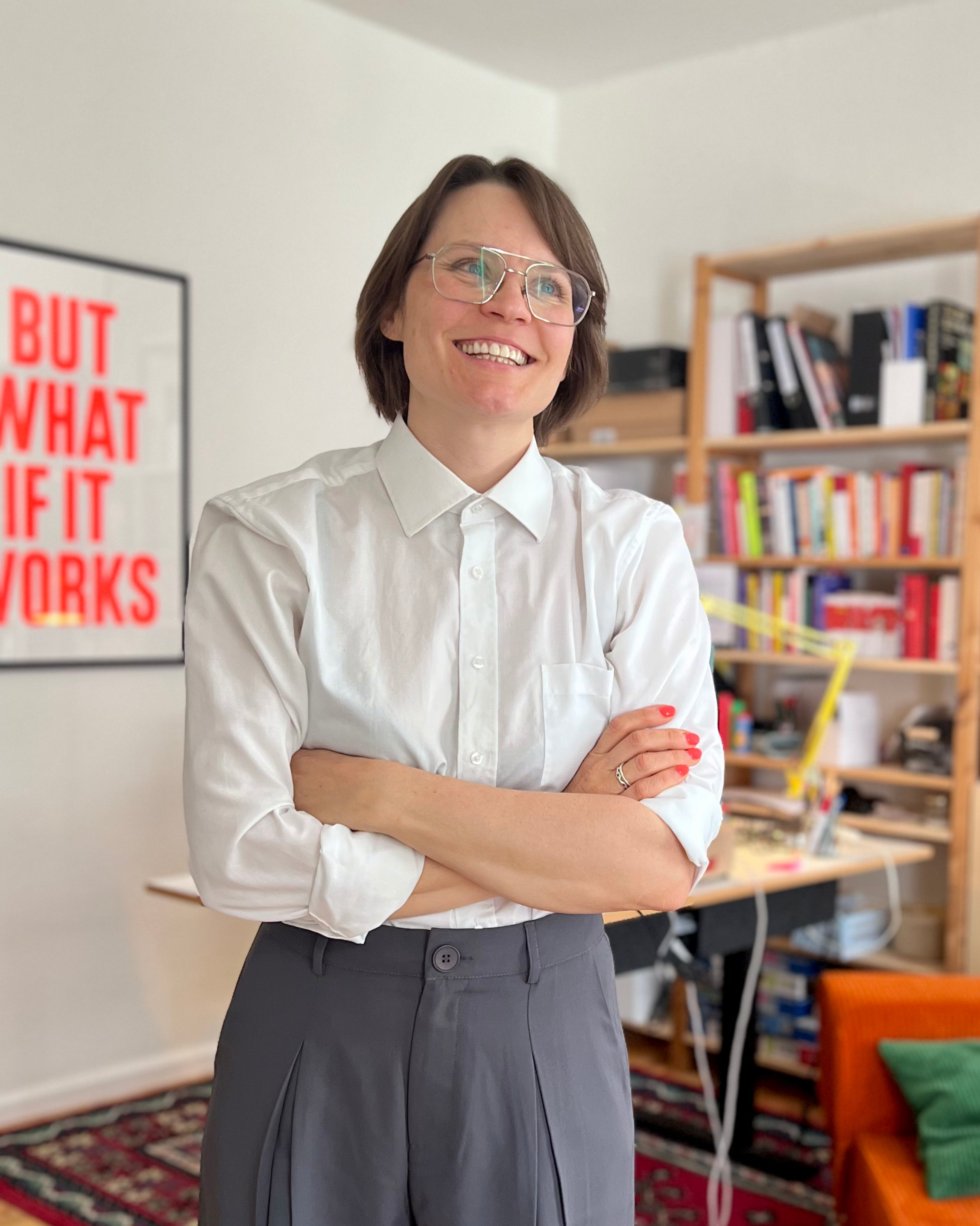 Eine Frau mit kurz braunen Haaren und Brille, die ein weißes Hemd und einen grauen Rock trägt, steht lachend mit verschränkten Armen in einem Raum mit Büchernregalen im Hintergrund.