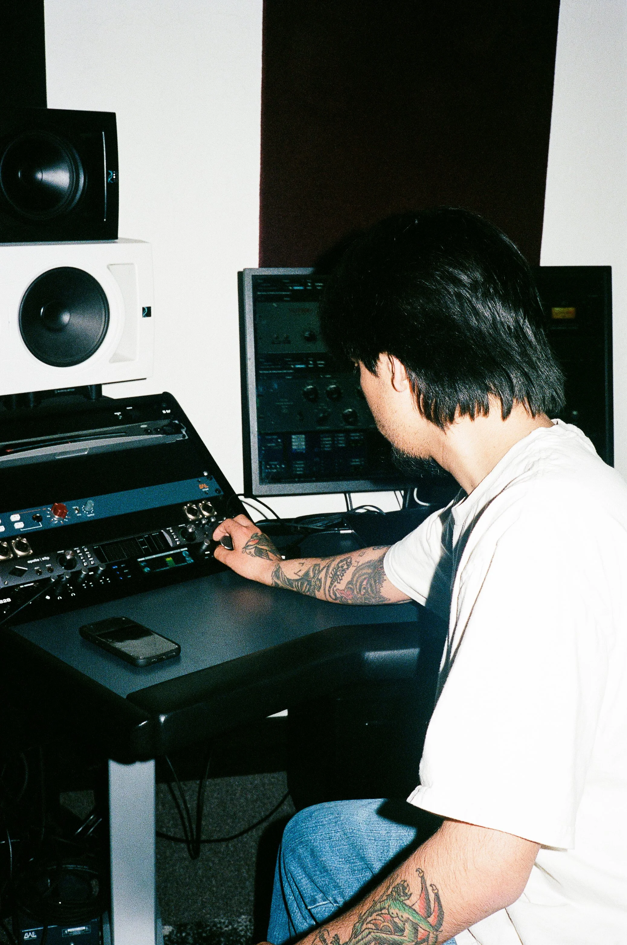 A man with black hair and tattoos on his arm using audio equipment in a recording studio. He is sitting at a mixing console with computer screens and speakers in the background.