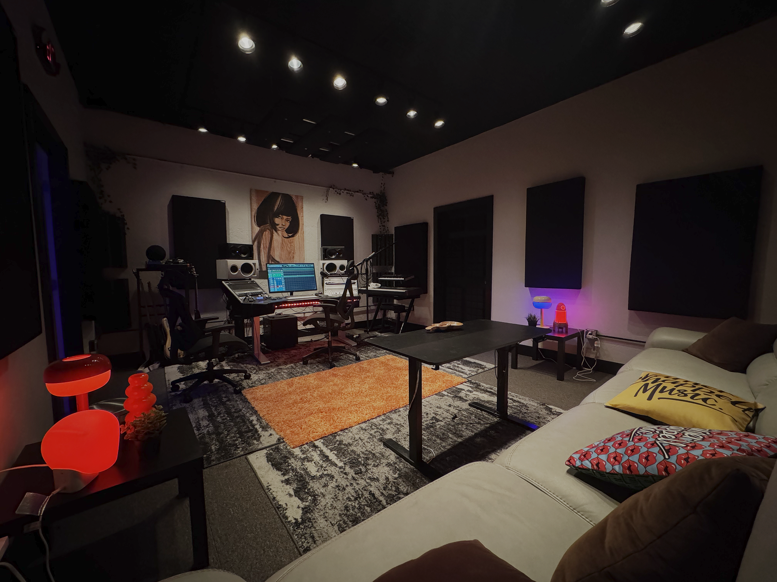 A music recording studio with a mixing console, speakers, a computer, and a microphone on a stand. There is a white sofa with pillows, a coffee table, and decorative lamps. The walls have soundproofing panels, and a painting of a woman with dark hair is hanging on the wall.