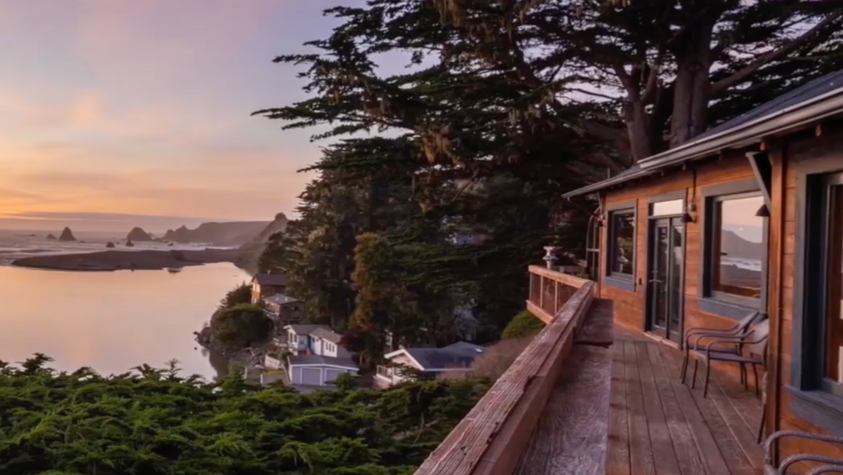 A scenic view from a wooden balcony overlooking a quiet bay with small houses, large trees, and rock formations in the ocean at sunset.