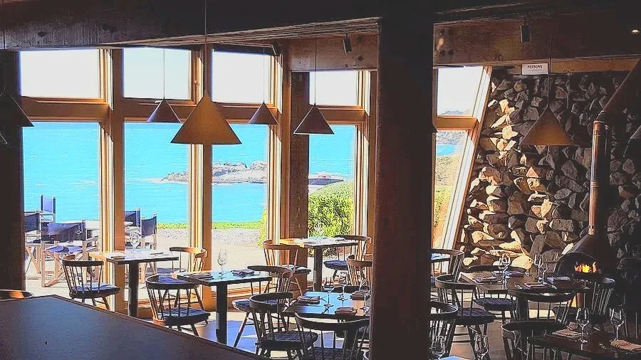 Interior of a restaurant with large windows overlooking the ocean, wooden tables and chairs, and a stone fireplace.