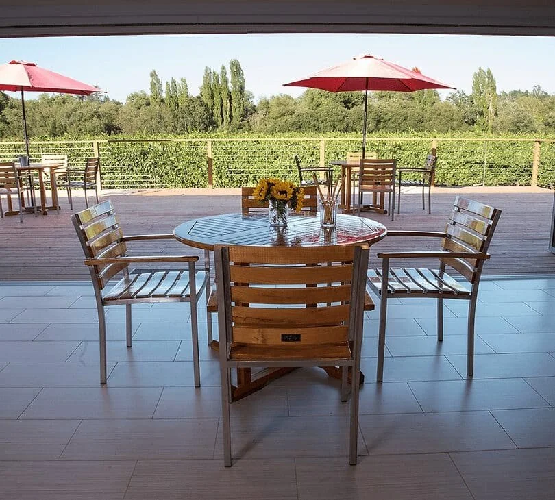 Indoor view of a dining table with four wooden chairs, a small vase with sunflowers, and two glass jars on it, overlooking a deck with additional chairs and umbrellas and a green landscape in the background.
