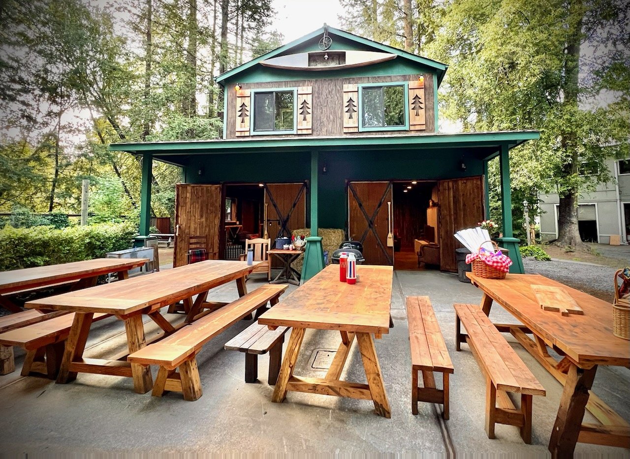 Outdoor backyard with wooden picnic tables, benches, and a two-story house with a green and brown exterior, surrounded by trees.