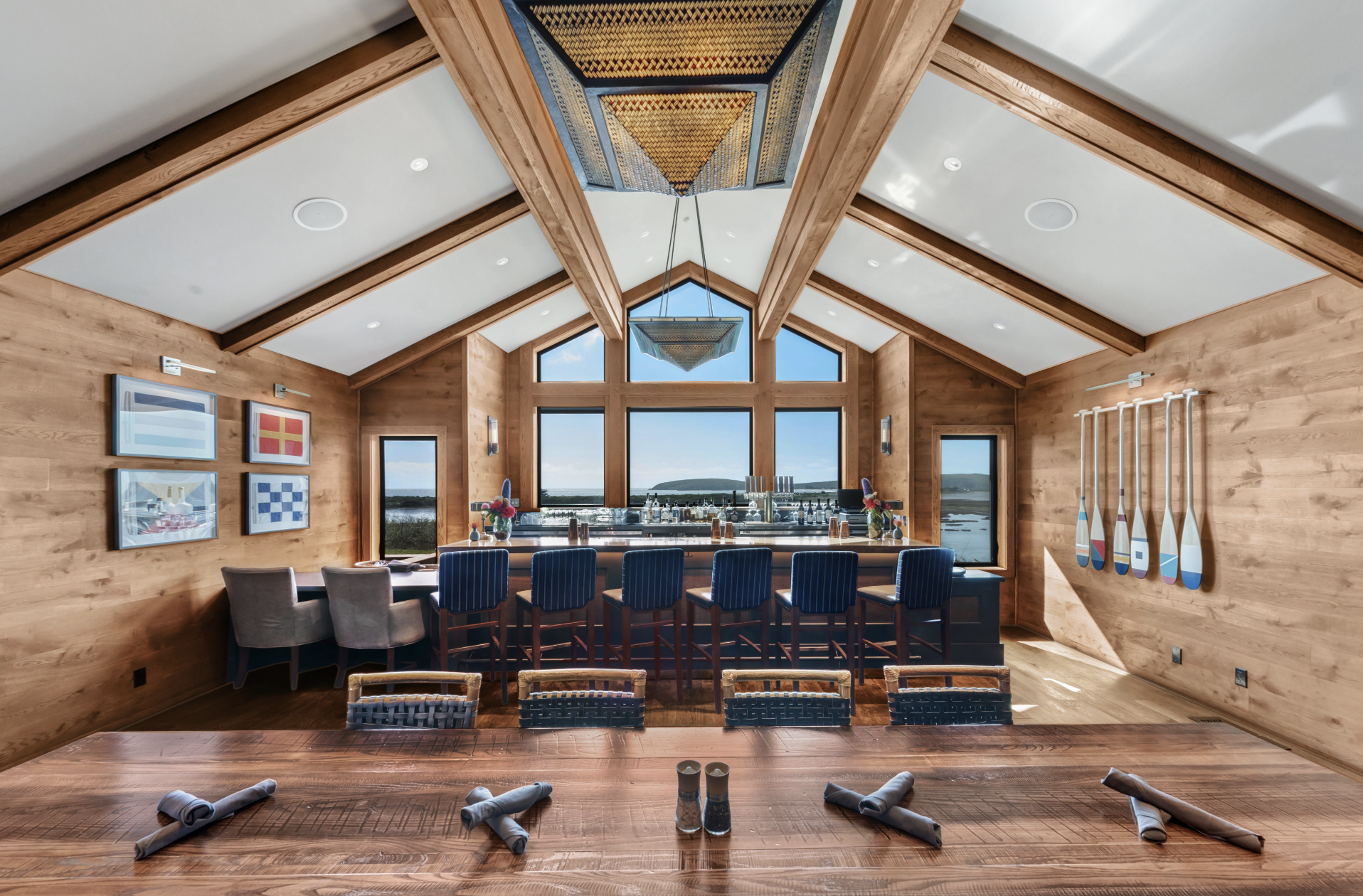 A spacious dining or lounge area with wooden walls, ceiling, and furniture. A row of blue high-back chairs lines a bar counter in front of large windows revealing a coastal landscape. There are four framed nautical art pieces on the wall and paddle decorations on the right wall. On the table in the foreground, three rolled napkins and a salt and pepper shaker set are visible.