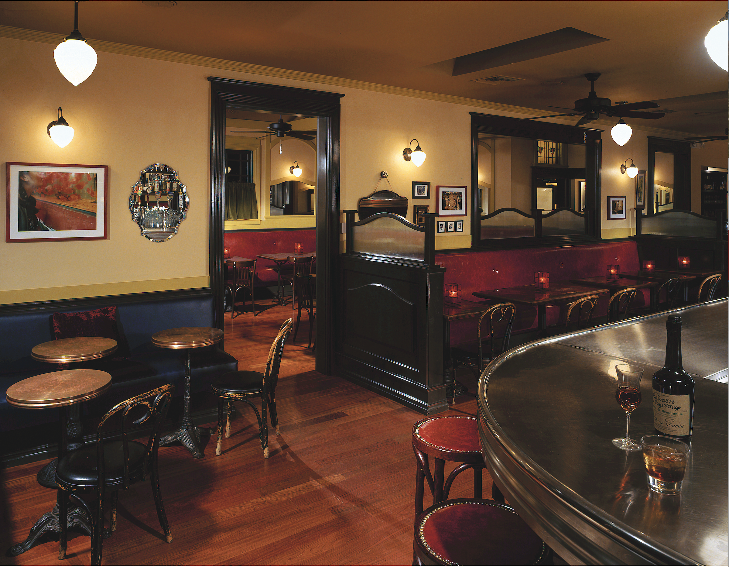 Interior of a cozy restaurant with wooden floors, yellow walls, black trim, and vintage-style light fixtures. There are small tables with chairs, a bar area with wine bottles, and framed artwork on the walls.