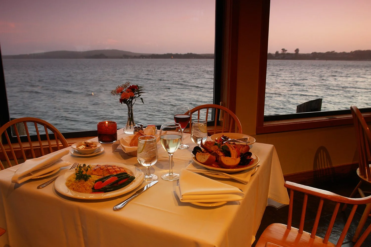 A dinner table set with plates of food, glasses of water and wine, a flower vase, and a candle, overlooking a lake at sunset.