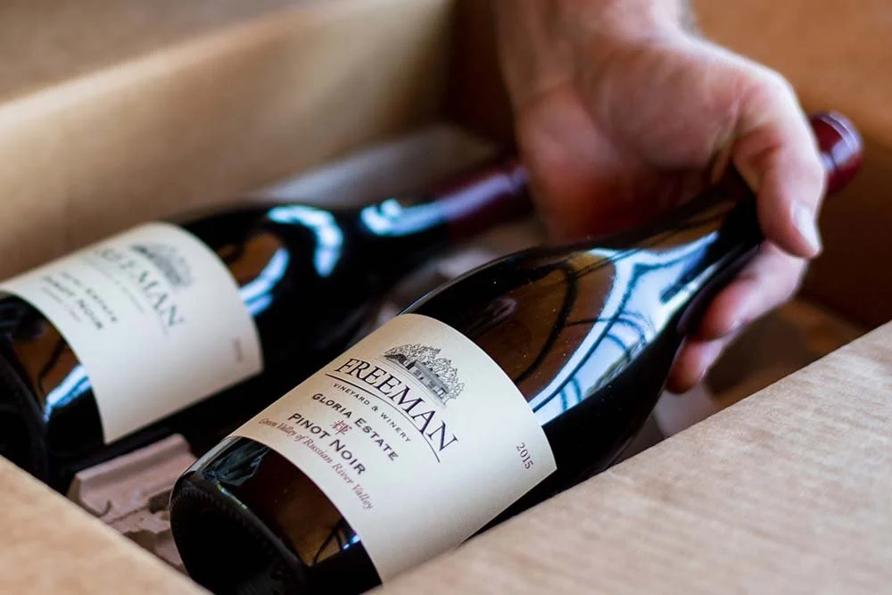 A person's hand lifting a bottle of Freemans Pinot Noir wine from a cardboard box.