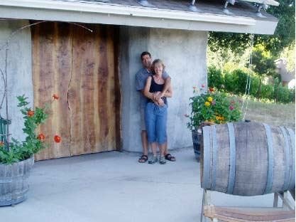 A couple stands close together outside near a concrete and wooden wall, with plants and a wooden barrel nearby.