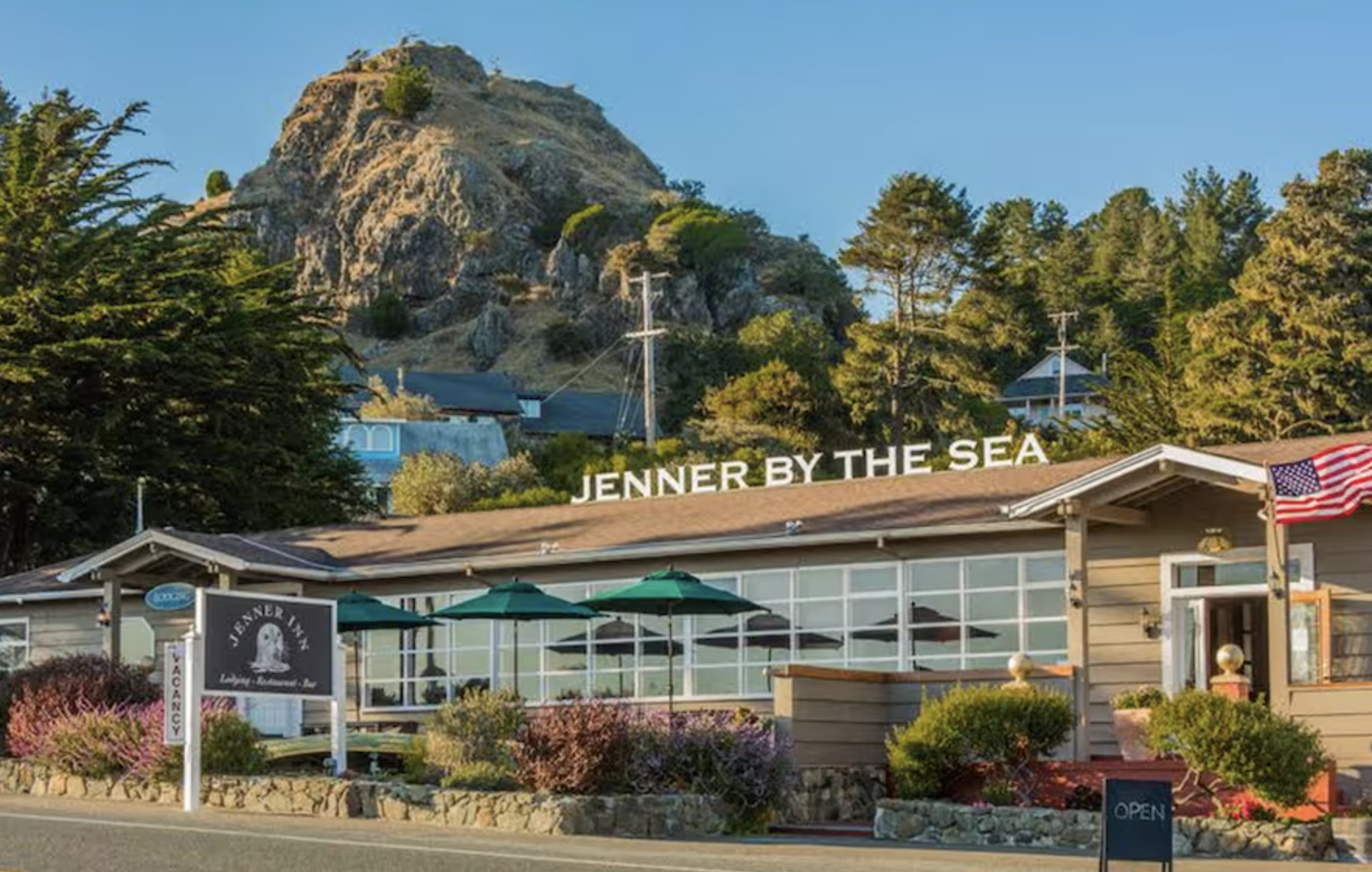 A seaside restaurant named Jenner Inn with outdoor patio seating, surrounded by greenery and flowers, with a mountain and trees in the background and a sign that says 'JENNER BY THE SEA' on the roof.