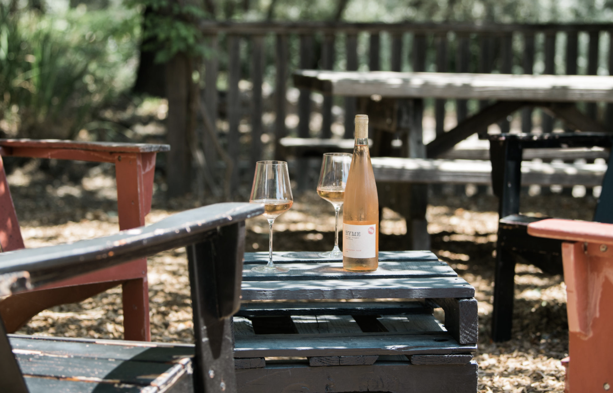 Outdoor scene with a small wooden table holding a wine bottle and two glasses of wine, surrounded by colorful chairs and a wooden deck in the background
