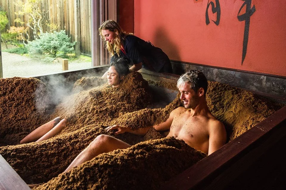 Two men and a woman relaxing in a traditional Japanese onsen bath filled with natural mineral spring mud, with a staff member helping one person.