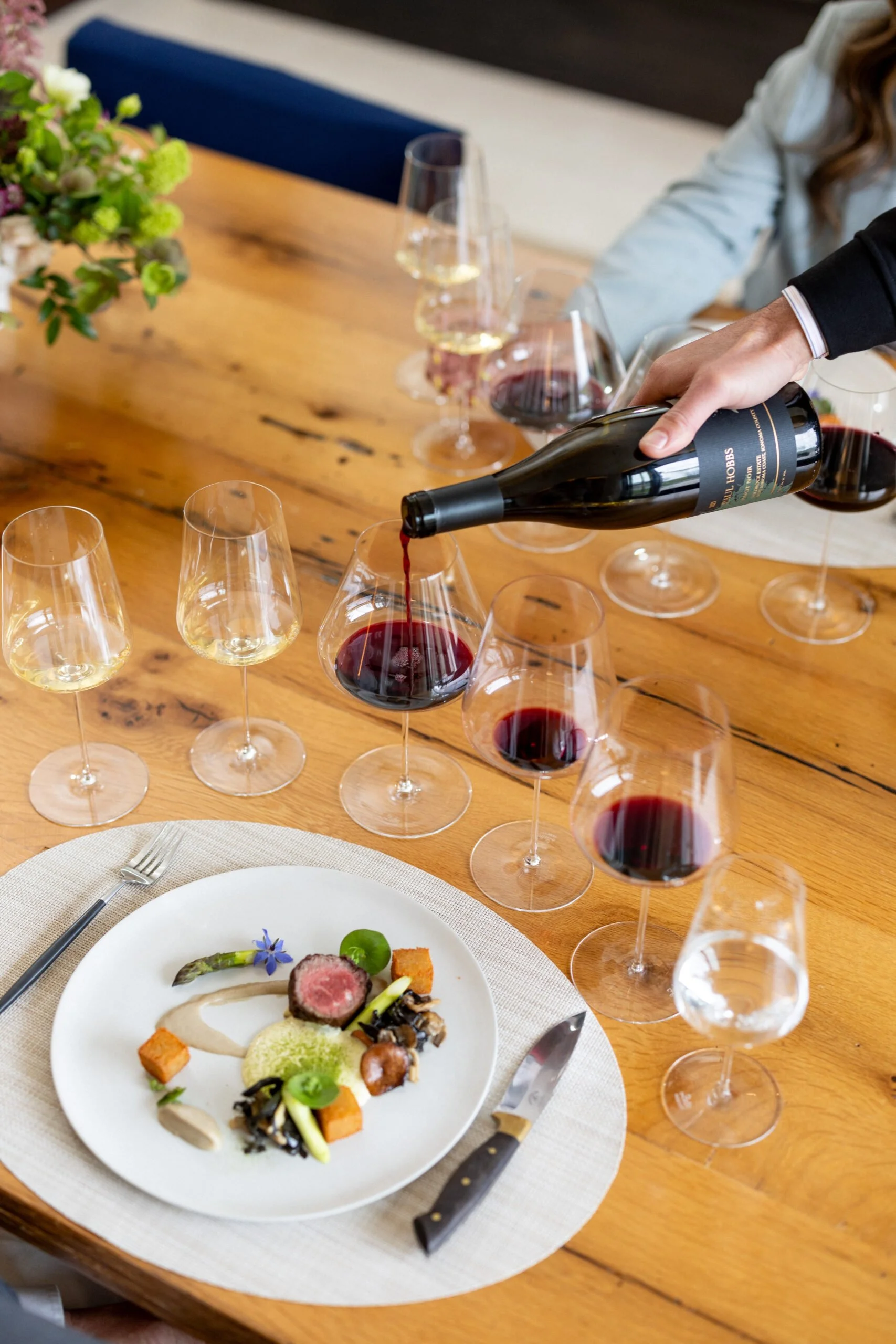 Person pouring red wine into a glass at a dinner table with multiple wine glasses, a plated dish, and a flower arrangement.