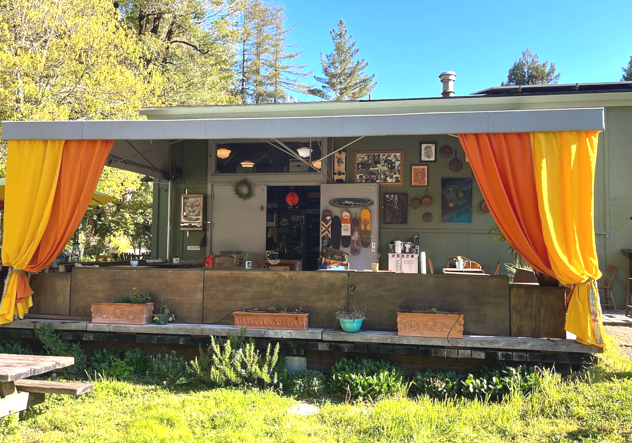 Outdoor stage with yellow and orange curtains, decorated with plants and art, set against trees and a blue sky.