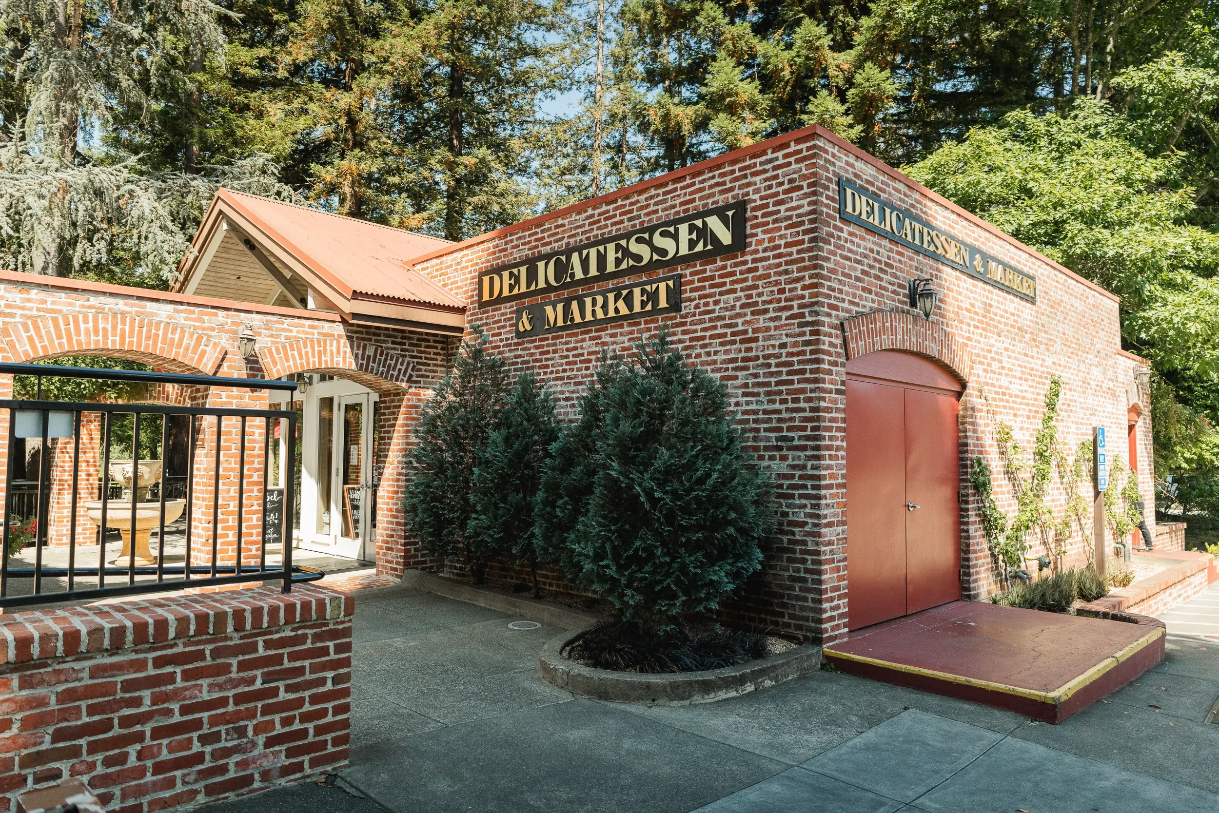 Exterior of a brick building with a sign reading 'Delicatessen & Market' and a small tree in front. The building has a reddish-brown door and large trees behind it.