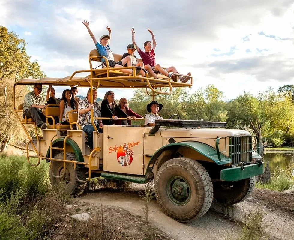 Group of people riding in an open-top safari vehicle with a sign that reads 'Safari West,' on a dirt road near a lake and trees with cloudy sky in the background.