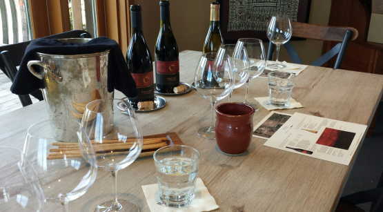 A wooden dining table set with wine bottles, empty wine glasses, water glasses, a clay pitcher, and a wine tasting menu.