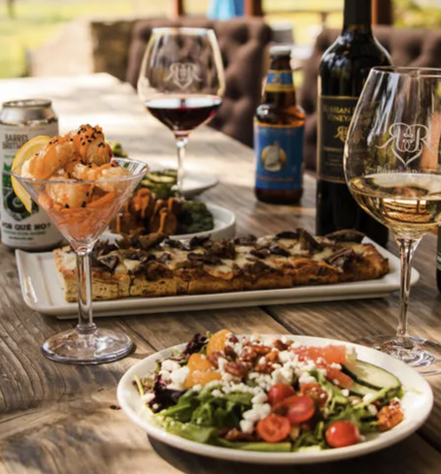 A table set for a meal outdoors with various food and drinks, including a salad, pizza, shrimp cocktail, red wine, beer, and a bottle of liquor.