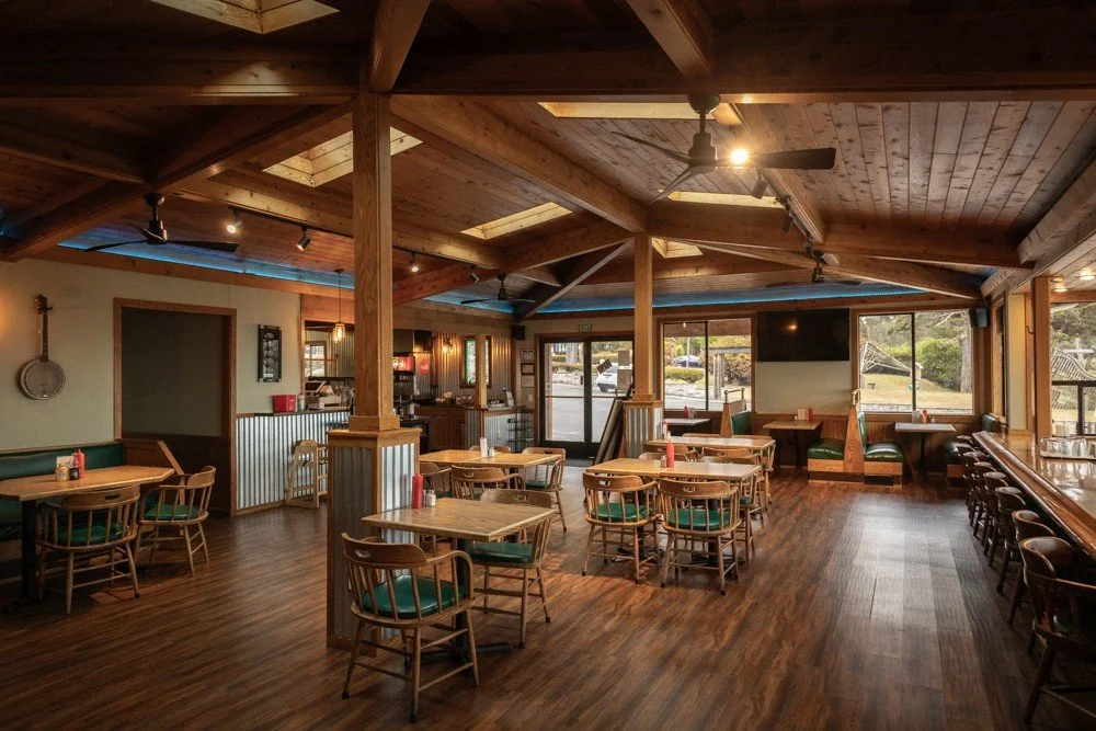 Interior of a rustic restaurant or cafe with wooden furniture, large windows, a TV, and ceiling fans.